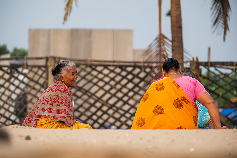 An older woman smiling toward two women in bright clothing on a sunlit street.
